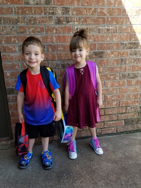 Two children standing in front of a brick wall with backpacks.