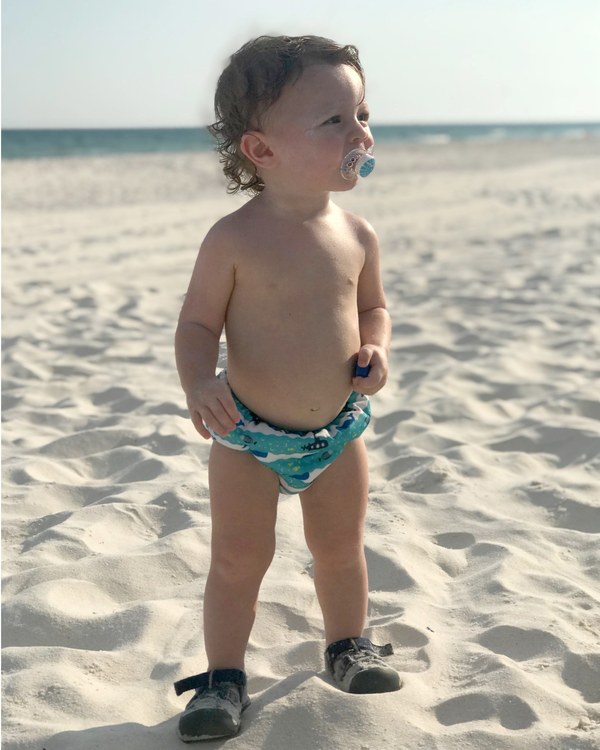 A baby standing on the beach with a pacifier in his mouth.