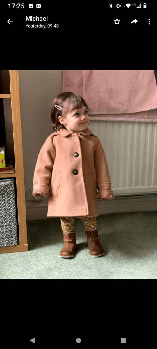 A little girl in a brown coat standing in front of a window.