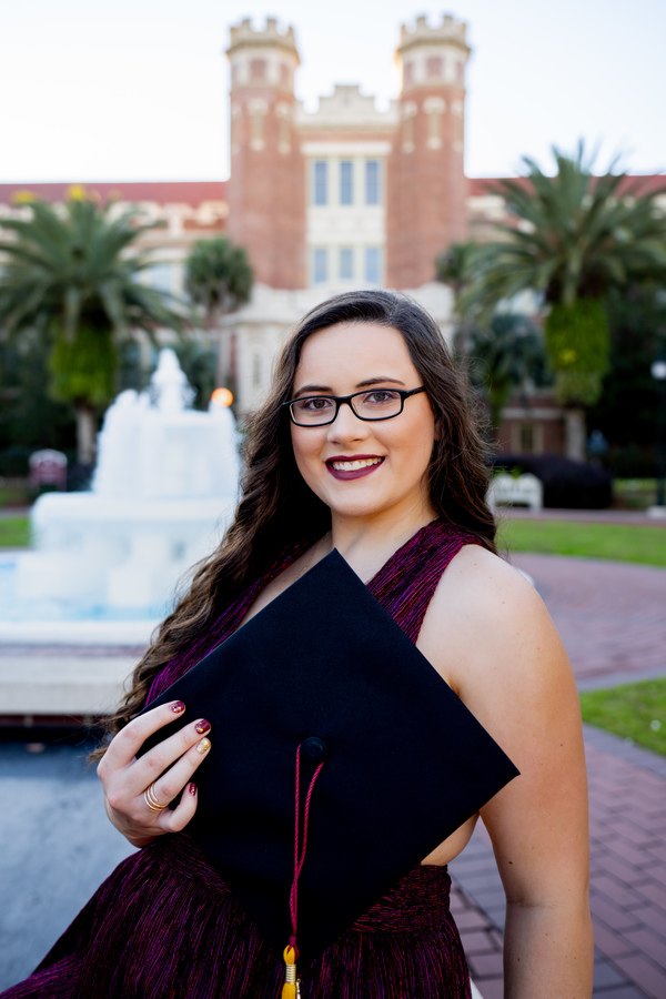A young woman holding her graduation cap in front of a fountain.