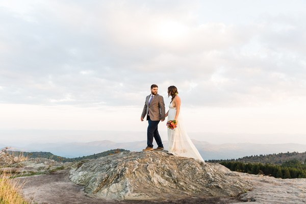 A bride and groom standing on top of a mountain at sunset.