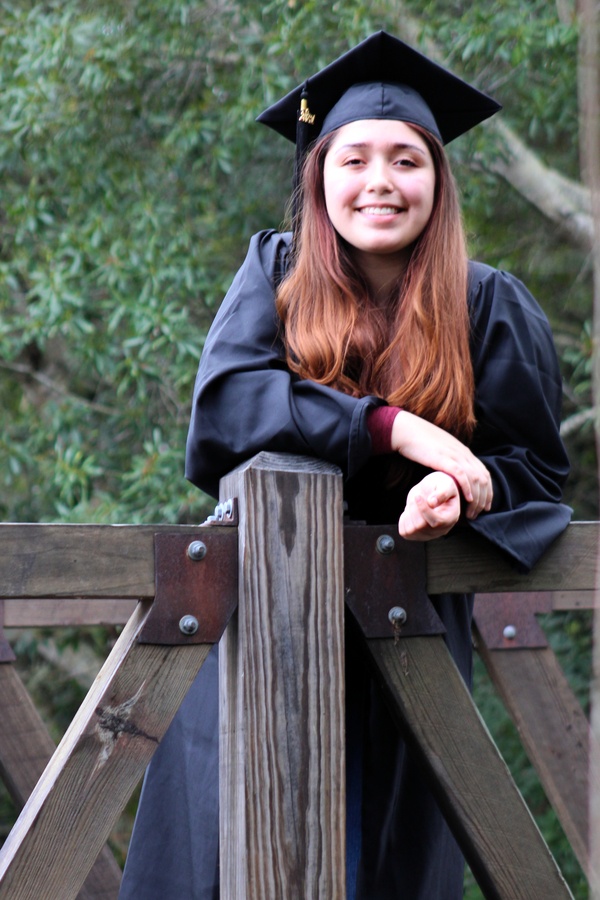 A woman in a graduation gown leaning on a wooden fence.