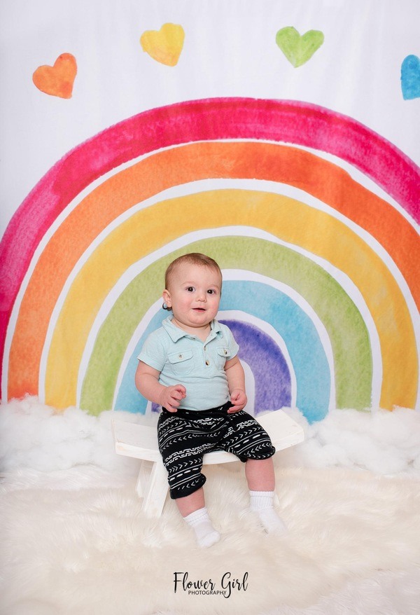 A baby sits on a bench in front of a rainbow backdrop.