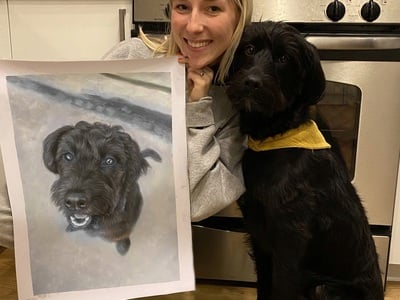A woman holding up a photo of a black and white oil painting of a dog, a beautiful transformation from picture to portrait painting.