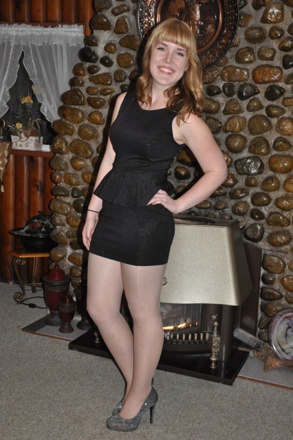 A young woman in a black dress posing in front of a fireplace.