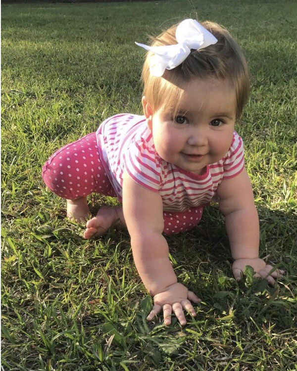 A baby crawling in the grass with a pink bow on her head.