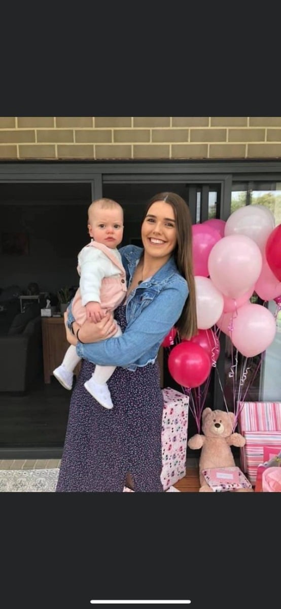 A woman holding a baby in front of pink balloons.